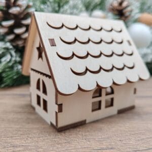 A miniature 3D laser cut wooden Christmas house ornament made from 3mm plywood, displayed on a wooden surface with a blurred winter background featuring pine cones and snowy trees.