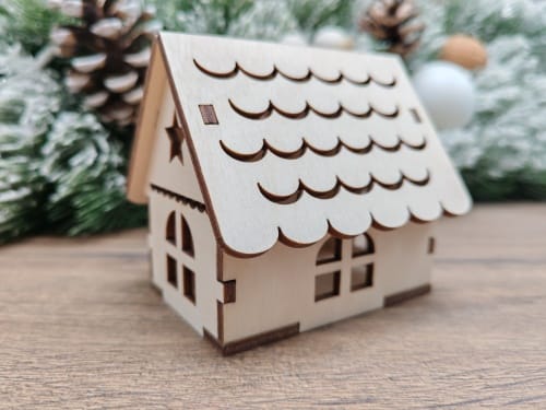 A miniature 3D laser cut wooden Christmas house ornament made from 3mm plywood, displayed on a wooden surface with a blurred winter background featuring pine cones and snowy trees.