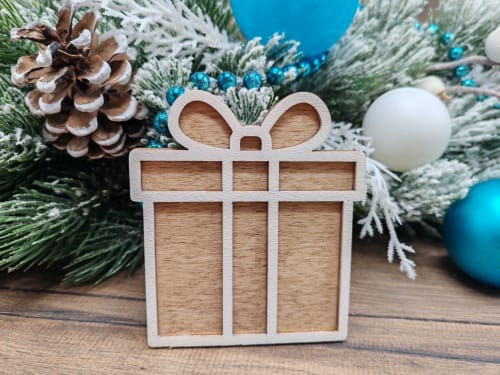Free 3D layered laser cut wooden Christmas present ornament in a gingerbread style, displayed as a finished craft project on a rustic wooden table with snowy holiday branches and decorations.