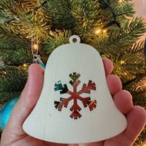 A hand holding a white laser-cut wooden Christmas ornament shaped like a bell with an intricate snowflake cut out in the center, showcased in front of a decorated Christmas tree.