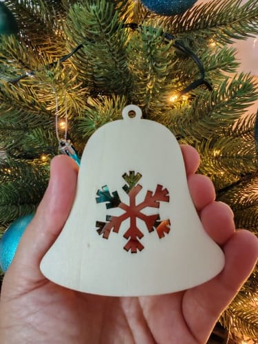 A hand holding a white laser-cut wooden Christmas ornament shaped like a bell with an intricate snowflake cut out in the center, showcased in front of a decorated Christmas tree.