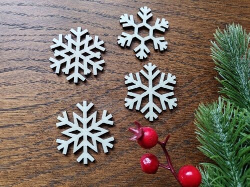 A set of four intricate wooden snowflake ornaments cut with a laser machine, placed on a dark wooden table next to green pine branches and red berries.